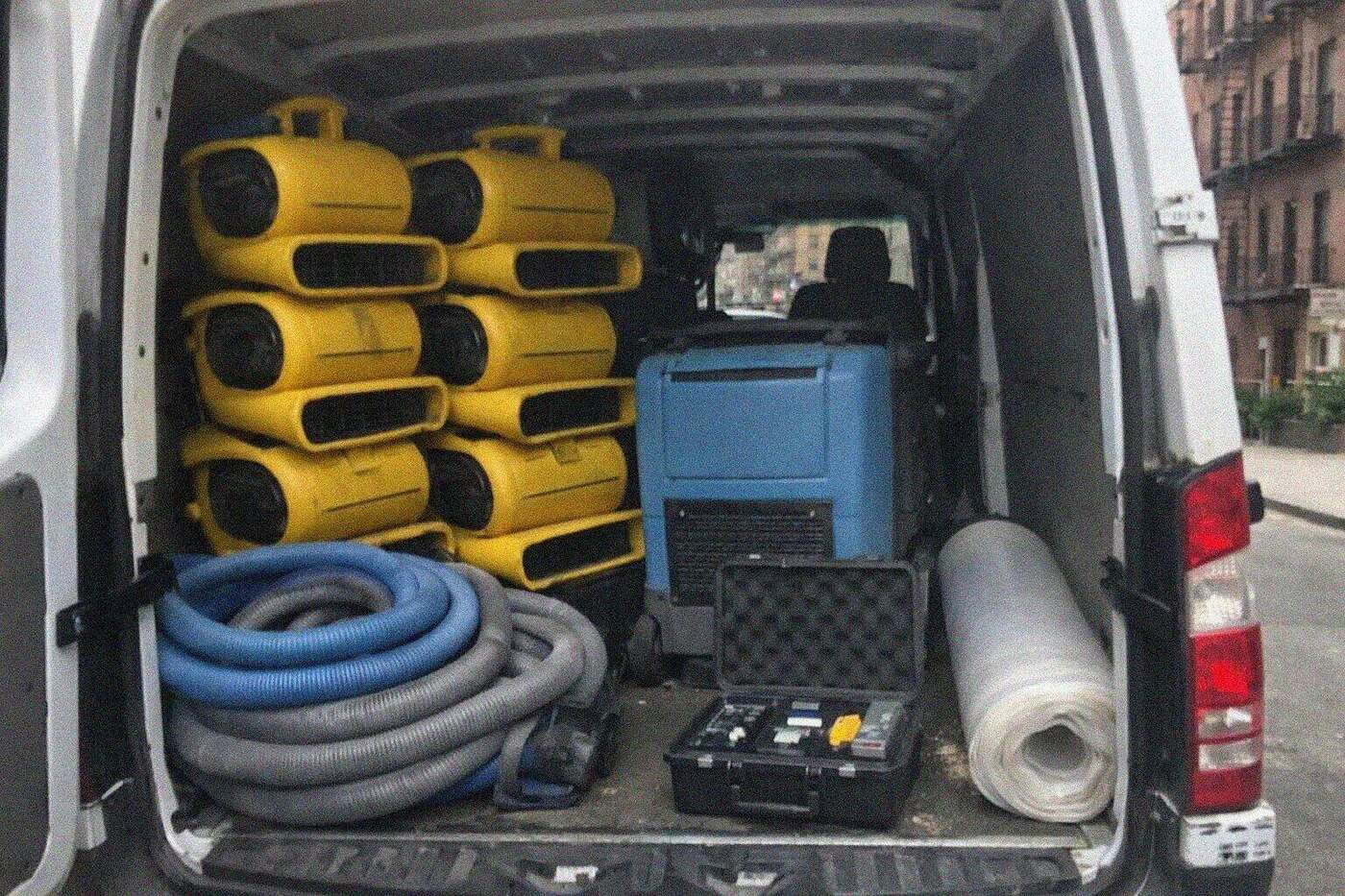 The open rear doors of a restoration van on a New York street, stacked with air movers, a dehumidifier and coiled hoses